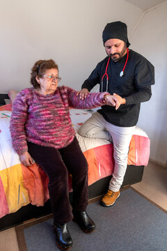 A male doctor on a house call checks on the health of an elderly woman sitting on her bed in her bedroom