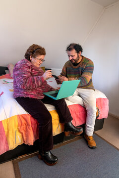 An adult man helps his elderly mother make bank transfers using her cell phone and a laptop