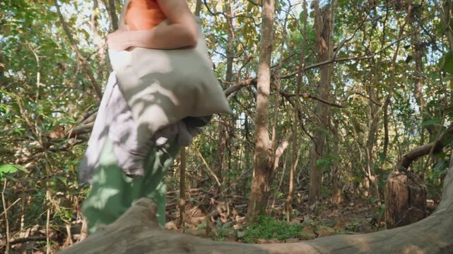 Close up of woman walking through dense forest carrying bags and wearing green pants. Woman holds various items including canvas tote bag plastic pouch with drawstring gray jacket draped