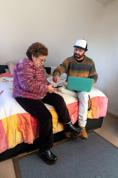 An adult man helps his elderly mother use her cell phone and a laptop