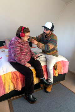 An adult man puts headphones on his mother's head so she can listen to her favorite music from his cell phone