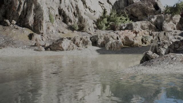 A rocky area with water at the forefront. The water reflects rocks and nearby plants. The location appears to be a natural outdoor setting during daytime.