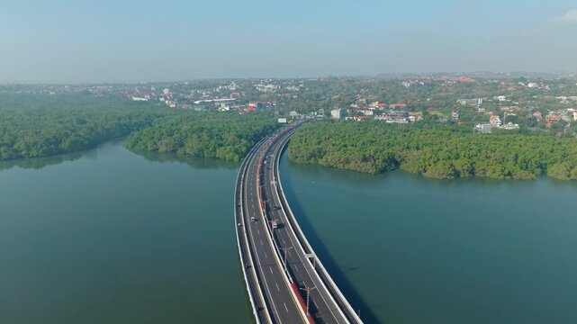 Aerial view of Bali Mandara Toll Road stretching across the water with lush mangrove forests and traffic in Kecamatan Kuta Selatan, Bali, Indonesia.