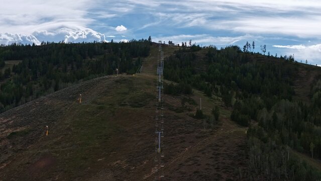 Aerial view of a ski lift ascending a mountain slope with pine trees and dry vegetation under a cloudy blue sky in Granby, Colorado, United States.