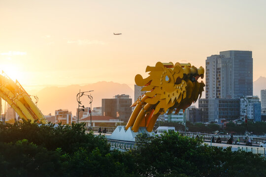 Yellow dragon structure adorns the bridge over the river during a bright afternoon with city buildings and lush green trees in the background view