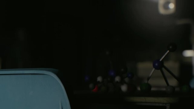 A molecular model sits on a table in a dark room. A projector casts light on the scene. The setting appears to be a laboratory or classroom focusing on science activities at night.
