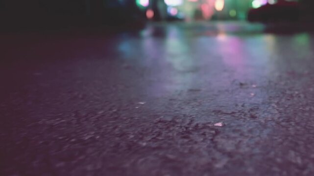 A view from the ground shows wet pavement during nighttime in a busy urban area. Bright lights can be seen reflecting off the surface. People may be nearby.