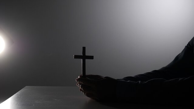 Person's hands holding a wooden cross against a dark background, reflecting concepts of faith, prayer, hope, and spiritual devotion in a somber and reverent atmosphere