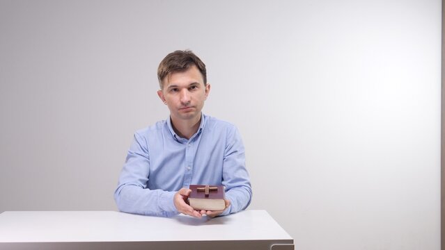 Man holding a bible with a wooden cross on its cover, seated at a table against a neutral background, conveying christian faith, devotion, prayerful study and spiritual reflection