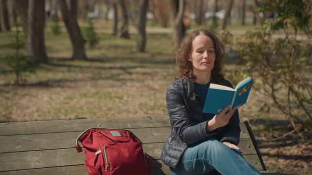 Close up of woman reading book on wooden bench in park with red backpack. Woman holds blue notebook occasionally turning pages while seated outdoors surrounded by trees and grass. Soft