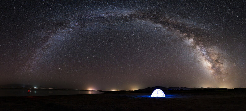 A vivid Milky Way arches over a lit tent in a dark, remote outdoor night landscape with faint distant lights on the horizon.