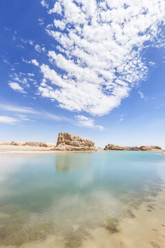 A calm clear lake sits among layered rock formations under a bright blue sky with scattered white clouds, in an arid landscape.