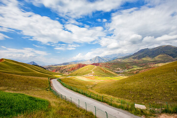 A winding paved road cuts through rolling grassy hills and colorful mountain valleys under a bright blue sky with scattered white clouds. © tai