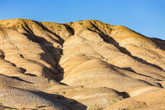 Layered eroded desert hills with distinct geological layers, lit by sunlight under a clear blue arid sky, showing natural terrain patterns.