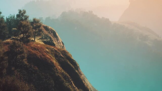 A rocky cliff stands tall with trees on top while soft sunlight illuminates the area at dawn. The foggy background adds depth to the landscape.
