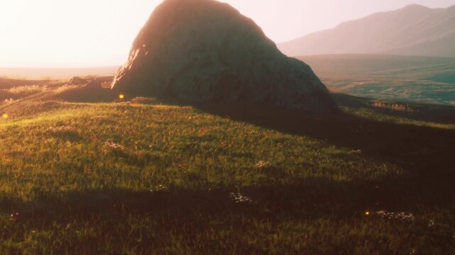 The sun sets over a grassy hill, highlighting a large rock. Mountains are visible in the distance, creating a scenic landscape. This view captures the end of the day with natural elements.