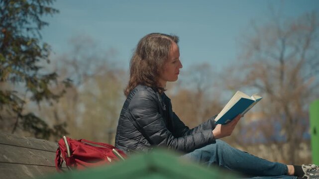 Close up of woman reading open book while seated on wooden steps outdoors with trees and sky in background. Woman wears dark jacket blue jeans sneakers red backpack rests beside her. Soft