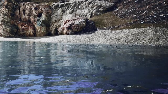 Water reflects the surrounding rocks and ground near a secluded shore. The scene is quiet with limited movement at the waters edge during daylight hours.