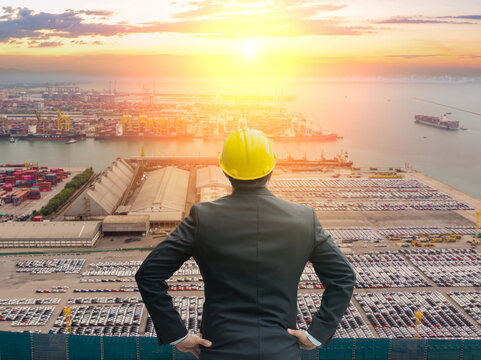 Businessman in hard hat looking over a car port and shipping terminal at sunset. Global trade concept. Human-created, not AI.