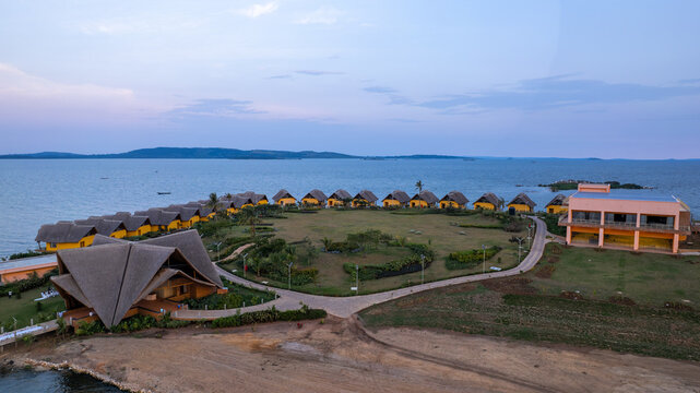 Kampala City, Uganda - 23 August 2026: Aerial view of Paradise Highland resort featuring thatched-roof bungalows along the lakeshore, a central lawn, and a modern building under a clear sky.