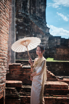 Portrait of Asian teenage girl dressed in traditional Thai outfit and umbrella with cheerful smile at old brick wall, summer holiday travel in Ayutthaya, ancient ruins heritage,  tourist destination.