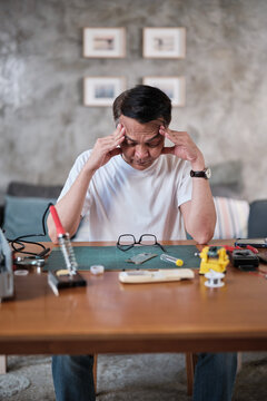 Mature Asian man concentrates on repairing home electrical appliance, disassembling and soldering circuit board, tired from precision detail focusing, taking a break, resting from his workshop hobby.