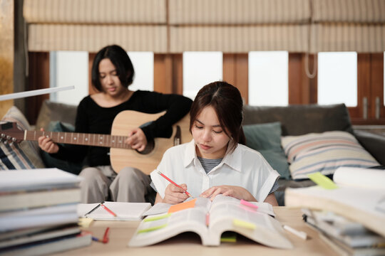 Two Asian teenage tutorials, girl concentrates on reading books for exam, learning at home, her friend relaxes by playing guitar music, self-study with textbooks, homeschooling together lifestyle.