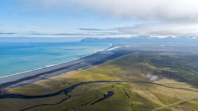 Aerial view of the dramatic coastline featuring black sand beaches, winding rivers, and vast green plains under a cloudy sky near Hellissandur, Snaefellsbaer, Iceland.