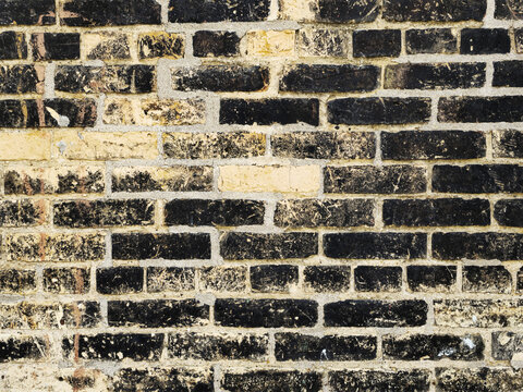 Close-up of an aged brick wall with dark brown and tan bricks arranged in a staggered pattern, showing weathered texture, mortar wear, and the rustic charm of historic masonry craftsmanship.