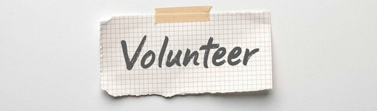 Call to Action: A handwritten note with the word Volunteer taped to a wall, serving as a simple yet powerful invitation to contribute time and effort to a cause.