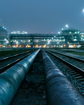 Large pipes run parallel along railway tracks. Industrial complex glows faintly behind the rails. Dark sky frames the mechanical expanse. Cold, dim lighting enhances the scene's isolation
