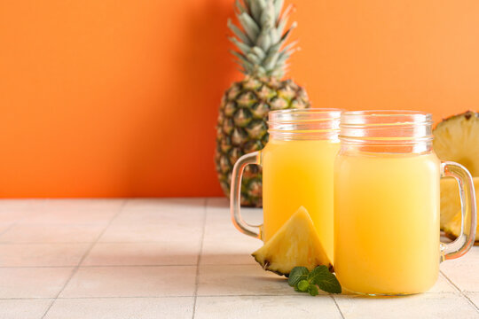 Mason jars of fresh pineapple juice with mint on white tile table against orange background