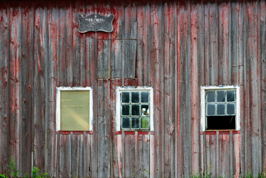 Weathered red barn wall with three windows framed in white, showing peeling paint, aged wood texture, and rustic charm, capturing the timeless character of rural architecture and farm heritage.