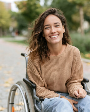 Warm smile radiates from woman in wheelchair. She wears cozy sweater and jeans, sitting calmly. Autumn leaves scatter around her on paved path. Trees softly frame the background with golden hues