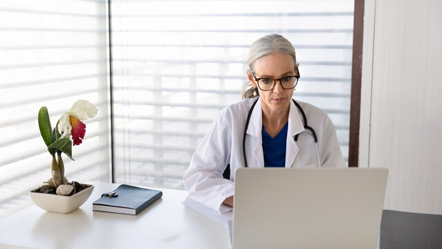 General practitioner working on laptop in clinic, consulting hospital patients, conducting telehealth appointments, reviewing treatment plans or test results, making administrative healthcare tasks