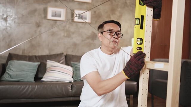 Asian mature man self-assembling wooden furniture with DIY tools, checking work precision with spirit&nbsp;level, home improvement, craft installation, hobby carpentry workshop, handyman active lifestyle.