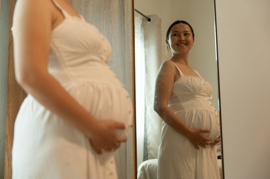 Close-up of a pregnant woman in a white button-front dress smiling toward the side, her reflection visible in the mirror.