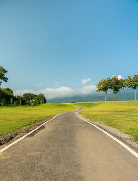 Winding country road curving through a green field toward misty mountains under a bright blue sky with soft clouds.