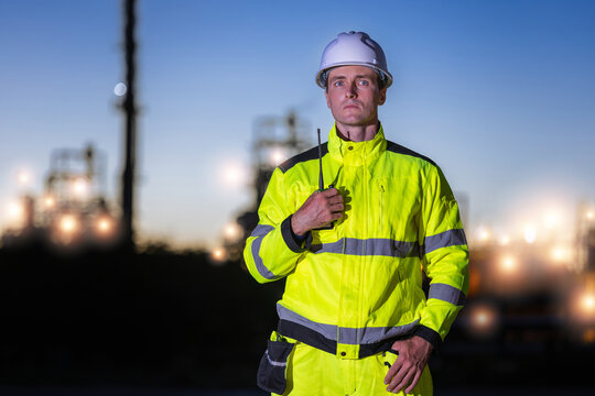 Industrial engineer holding walkie talkie at oil refinery during twilight, petroleum plant worker standing at facility at night, energy industry professional with radio at industrial plant evening
