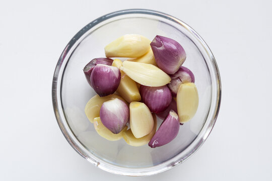 Top view of peeled garlic cloves and red shallots mixed in a small glass bowl on a white background.
