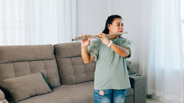A female flutist in a green shirt sits gracefully on gray sofa, playing a silver flute. The cozy living room setting adds warmth and serenity, showcasing her passion for music in a casual environment