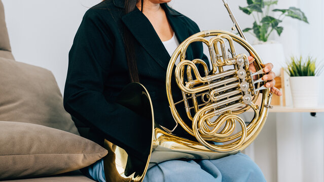 A woman sits on a cozy sofa in her living room, playing a shiny gold French horn. The warm lighting and serene atmosphere reflect her passion for music