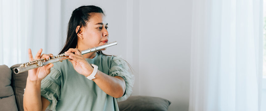 A female flutist in a green shirt sits gracefully on gray sofa, playing a silver flute. The cozy living room setting adds warmth and serenity, showcasing her passion for music in a casual environment