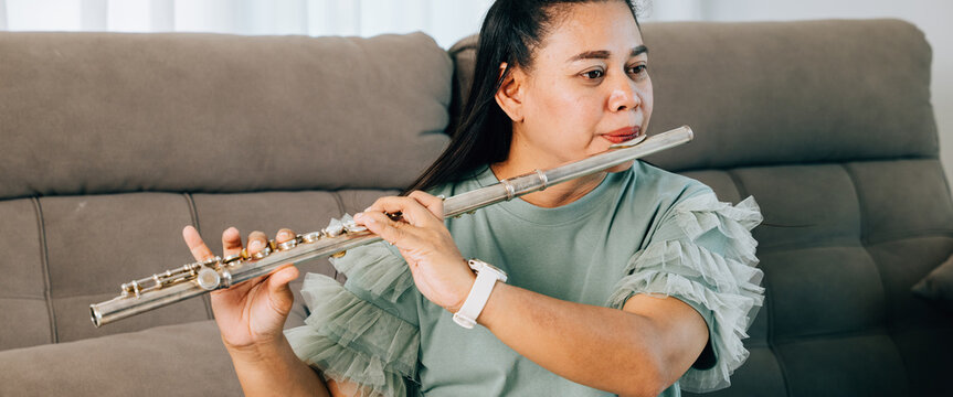 A female flutist in a green shirt sits gracefully on gray sofa, playing a silver flute. The cozy living room setting adds warmth and serenity, showcasing her passion for music in a casual environment