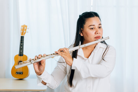 A flutist in a white shirt sits on a chair at home, blowing into her silver flute. The living room curtain backdrop accentuates