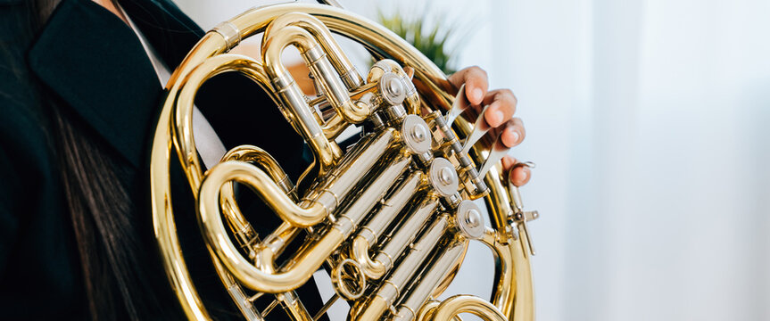 A woman sits on a cozy sofa in her living room, playing a shiny gold French horn. The warm lighting and serene atmosphere reflect her passion for music