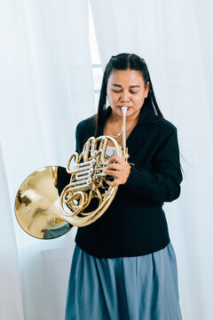 A woman stands at home in front of elegant curtains, playing a shiny gold horn. The warm lighting and her graceful demeanor bring out the beauty of her musical