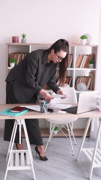 Vertical video. Corporate lifestyle. Employee routine. Woman specialist drinking coffee analyzing business document at office desk with laptop.
