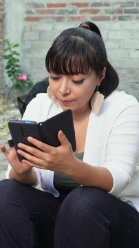 Woman sitting in her home patio checking her cellphone while her dog walks behind her