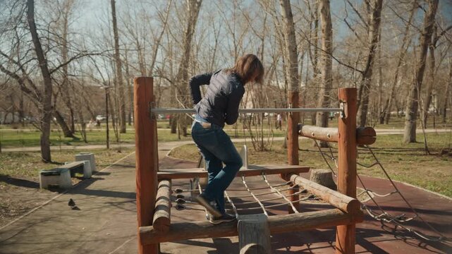 Woman navigates wooden playground structure in outdoor park performing physical activity on monkey bars and ropes. Woman wears casual clothing including jeans as woman moves through play